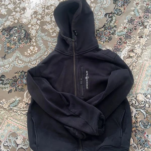  Äkta Svart hoodiejacka från Sail Racing - Svart hoodiejacka från Sail Racing med dragkedja framtill och på bröstfickan. Jackan har en stor huva och är tillverkad i mjukt syntetmaterial. Snyggt broderad logga på bröstet och praktiska fickor på sidorna. Perfekt för chill dagar och streetwear.