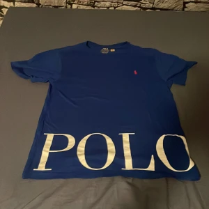 Blå Polo Ralph Lauren t-shirt - Snygg blå t-shirt från Polo Ralph Lauren med stor vit POLO-text längst ner och klassisk röd logga på bröstet. Modellen är kortärmad och har en normal passform. Tillverkad i mjuk bomull som känns skön mot huden. Perfekt för dig som gillar stilrena och ikoniska plagg.