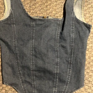 Mörk jeanskorsett med dragkedja - Snygg mörkblå korsettopp i jeansmaterial med guldfärgad dragkedja framtill. Modellen är croppad och har breda axelband. Materialet är en mix av bomull, polyester och elastan för en bekväm och tight passform. Perfekt för att ge din outfit en cool edge.