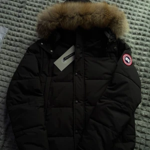 Svart dunjacka med päls från Canada Goose - Säljer en svart dunjacka från Canada Goose med äkta päls på huvan. Jackan har klassisk patch på ärmen, dragkedja och knappar framtill samt flera praktiska fickor. Perfekt för kalla dagar.