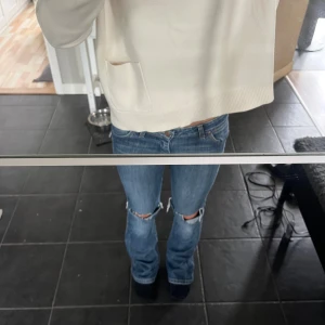 Blå bootcut jeans med slitna knän - Säljer ett par blå jeans från Wrangler med låg midja och bootcut passform. Jeansen har slitna hål vid knäna och klassiska fem fickor. Materialet är jeans och de har en snygg tvättad look med ljusare partier. Perfekt för dig som gillar en avslappnad och trendig stil. Midja 38cm rakt över, innerbenslängd 82