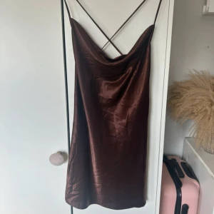 Brun satin klänning med smala axelband - Supersnygg brun klänning i satin med smala axelband som korsas i ryggen. Klänningen har en draperad halsringning och är kort i modellen, perfekt för dig som gillar en chic och stilren look. Materialet är glansigt och mjukt, vilket ger en lyxig känsla.