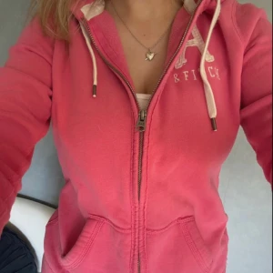 Rosa zip up hoodie från Abercrombie & Fitch - Säljer en snygg rosa hoodie från Abercrombie & Fitch med dragkedja och vita snören. Tröjan har huva och en broderad logga på bröstet. Perfekt för chill dagar och enkel att matcha med jeans eller leggings.