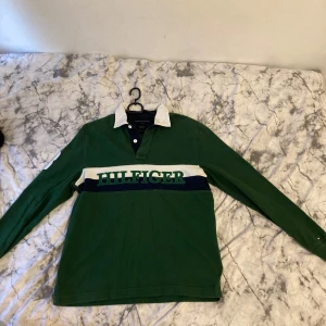 Grön pikétröja Tommy Hilfiger M - Säljer en grön långärmad pikétröja från Tommy Hilfiger i klassisk fit. Tröjan har vit krage, blå och vit rand över bröstet med stor Hilfiger-text, samt en vit siffra på ärmen. Perfekt för dig som gillar sportig och preppy stil.