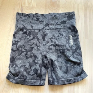 Grå camo shorts från Gymshark - Snygga grå träningsshorts från Gymshark med camouflagemönster och hög midja. Säljer vidare pga kommer inte till användning. Storlek M men sitter bra på mig som är xs/s