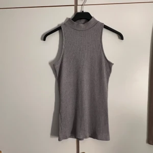 Grå ribbad ärmlös topp 157 - Säljer en grå ribbad ärmlös topp från 157 med hög hals. Toppen har en slim passform och är tillverkad i mjukt, stretchigt material som sitter snyggt på kroppen. Perfekt att styla med jeans eller kjol för en clean look.