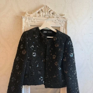 Svart paljettjacka från Zara - Snygg svart jacka från Zara med paljettdetaljer över hela ytan. Jackan är croppad och har lång ärm, perfekt för att lyfta din outfit. Materialet är strukturerat och ger en exklusiv känsla. Stängs framtill utan synliga knappar eller dragkedja. Endast testad 🥰