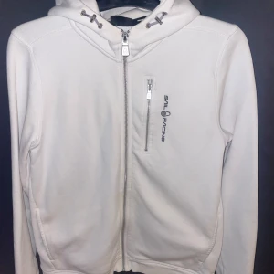 Vit hoodie från Sail Racing med dragkedja - Säljer en fräsch vit hoodie från Sail Racing med hel dragkedja framtill och en extra zip-ficka på bröstet. Hoodien har snörning i huvan och diskret logga på både bröstet och huvan. Perfekt för chill eller när du vill ha en clean look.