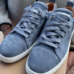 Blå mocka sneakers från Louboutin - Snygga blå sneakers i mocka från Christian Louboutin med klassisk röd sula och vita kanter. Skorna har rund tå, platt sula och matchande blå snörning. Insidan är i beige skinn för extra komfort. Perfekta för dig som vill sticka ut med exklusiv stil. Och har använt dem 1 gång men den är fortfarande i samma skick