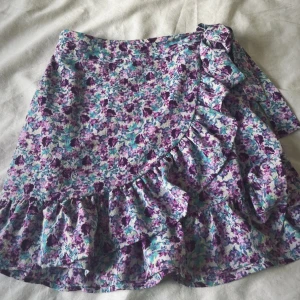 Blommig omlottkjol med volang - Superfin kort omlottkjol med blommigt mönster i lila och blått. Kjolen har volangdetaljer längs nederkanten och omlottknäppning med knyt i sidan. Aldrig använt så inga skavanker. Köpt på newyorker för runt 200kr 