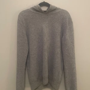 Grå cashmere hoodie -  grå stickad cashmere hoodie. Tröjan har en enkel och clean look, perfekt för dig som gillar en avslappnad stil. Materialet är mjukt och känns skönt mot huden. Passar dig som vill ha en basic men ändå snygg tröja.