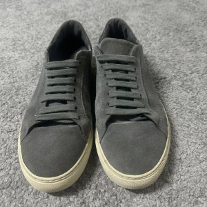 Grå mockasneakers från Axel Arigato - Snygga sneakers i grå mocka från Axel Arigato med klassisk låg profil och vit sula. Skorna har rund tå, snörning och diskret branding på sulan. Perfekta för en clean och stilren look. Riktigt fräscha. Hör av er om ni undrar något! BVSA