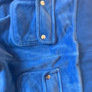 Blå velourbyxor Juicy Couture XS - Blåa mjukisbyxor från Juicy Couture i velour med bred resår och snörning i midjan. Byxorna har raka ben, stora fickor framtill med snygga metalldetaljer och broderad JC-logga på ena benet. Perfekta för en chill och trendig look.