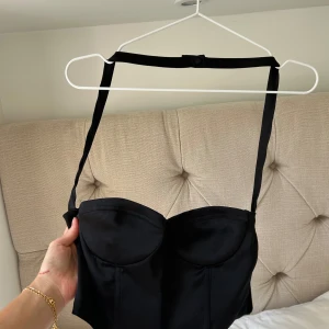 Svart korsett-topp med halterneck - Säljer en svart korsett-topp med snygg halterneck och croppad passform. Toppen har vadderade kupor och smala band som korsas över bröstet och knäpps i nacken. Perfekt att styla med höga byxor för en trendig look.