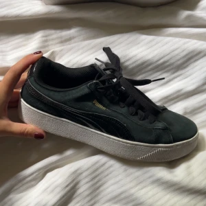 Svarta Puma sneakers i mocka - Snygga svarta Puma sneakers i mocka med klassisk vit sula och svarta snören. Skon har den ikoniska Puma-loggan i guld på sidan och svart läderdetalj längs sidan. Perfekt för dig som gillar stilrena och sportiga skor.