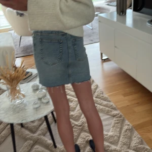 Blå jeanskjol med rå kant - Trendig blå jeanskjol med rå, fransig kant nertill och klassiska bakfickor. Kjolen har en kort passform och cool asymmetrisk knappstängning framtill. Perfekt för dig som gillar en avslappnad och edgy stil.