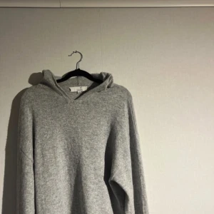 Grå hoodie i kashmir från Carmés - Mjuk och lyxig grå hoodie från Carmés i kashmir. Tröjan har huva, lång ärm och en clean look utan tryck eller detaljer. Perfekt för dig som gillar en chill och stilren vibe. Säljes i originalkartong.