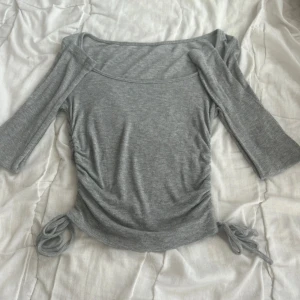 Grå ribbad topp med knyt i sidan - Superfin grå ribbad topp med trekvartsärm och snygga knytband i sidorna.Rynkade detaljer längs sidorna för en cool look. Perfekt att styla med jeans eller kjol för en trendig outfit.
