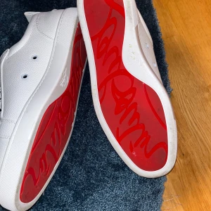 Vita Louboutin skor  - Exklusiva vita sneakers från Christian Louboutin i präglat skinn med ikonisk röd sula och guldfärgad logga på innersulan. Klassisk låg modell med snörning och rund tå, perfekt för dig som vill sticka ut med stilrena detaljer.