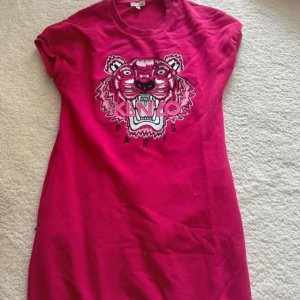 Rosa Kenzo klänning med tigertryck - Cool rosa kortklänning från Kenzo med ikoniskt tigerbroderi och texten 'Kenzo Paris' framtill. Klänningen har korta ärmar och är gjord i mjuk bomull, perfekt för en avslappnad och trendig look som passar både fest och vardag. Diskreta fickor på sidan
