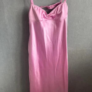 Rosa långklänning med öppen rygg - Snygg rosa långklänning i satinliknande material med smala axelband och öppen rygg. Klänningen har en lätt glansig finish och en figurnära passform som ger en elegant siluett. Perfekt för dig som vill sticka ut med en feminin look.