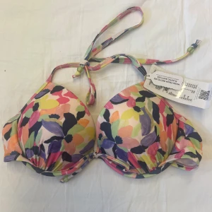 Färgglad blommig bikini från Together - Snygg bikiniöverdel från Together med push up och plunge-modell. Färgglatt blommigt mönster i rosa, lila, gul, grön, blå och svart. Knytband i nacken och under bysten. Tillverkad i återvunnen polyester för en hållbar stil på stranden.