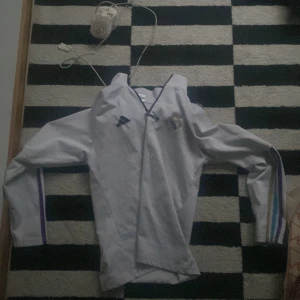 Real Madrid långärmad fotbollströja - Säljer en långärmad Real Madrid fotbollströja från Adidas i vitt med klubbens emblem på bröstet. Tröjan har klassiska Adidas-detaljer och färgade ränder längs ärmarna i lila, blått och gult. Perfekt för dig som älskar fotboll och vill visa ditt stöd för Real.