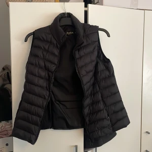Svart dunväst från Softy Collection - Svart dunväst från Softy Collection i storlek XL. Västen har quiltad design, hög krage och dragkedja framtill. Perfekt att slänga över en hoodie för extra värme. Snygg och enkel att matcha med det mesta.
