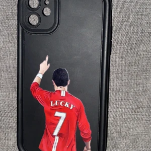 Svart mobilskal med fotbollstryck iPhone 11 - Snyggt svart mobilskal till iPhone 11 med motiv av en fotbollsspelare i röd tröja med texten 'LUCKY' och nummer 7 på ryggen. Skalet har en sportig vibe och är gjort i tålig plast som skyddar mobilen. Perfekt för dig som gillar fotboll och vill ha en unik look på mobilen.