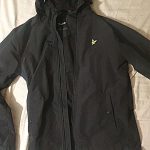 Svart vindjacka från Lyle & Scott - Svart vindjacka från Lyle & Scott med huva och dragkedja framtill. Jackan har den klassiska gula loggan på bröstet och praktiska fickor med knappar. Perfekt för blåsiga dagar och enkel att matcha med streetwear eller casual outfits.
