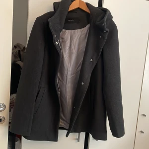 Svart kappa med huva från Vero Moda - Stilren svart kappa från Vero Moda i storlek M. Jackan har huva, dragkedja och knappar framtill samt två fickor. Fodrad insida för extra komfort. Perfekt för kyliga dagar när du vill hålla stilen enkel och clean.