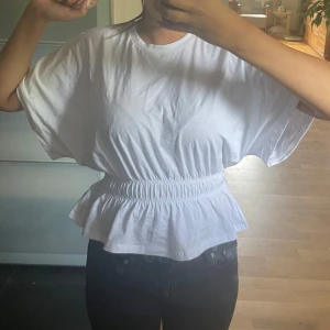 Vit blus med markerad midja - Säljer en vit blus med korta ärmar och rund hals. Toppen har en snygg smockad midja som ger en peplum-effekt och gör att den sitter superfint. Perfekt till jeans eller kjol för en fräsch look. Materialet känns mjukt och luftigt.