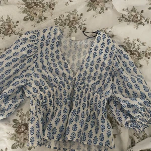 Blåvit blus med v-ringning, stl S - Supersöt blus i vitt med blått mönster, v-ringad och med smockad midja. Långa puffärmar med resår vid ärmslut. Perfekt för dig som gillar romantisk och trendig stil. Luftigt bomullsmaterial som passar till både jeans och kjol.