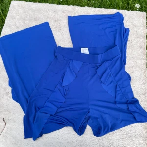 Blå vida byxor med volang Gina Tricot - Snygga blå byxor från Gina Tricot i mjukt polyestertyg med elastan. Vida ben och dekorativa volanger längs framsidan ger en trendig look. Perfekta för dig som gillar att sticka ut med färg och detaljer.