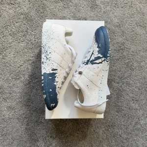 Maison Margiela sneakers med färgstänk - Unika sneakers från Maison Margiela i vitt tyg med coola mörkblå färgstänk över hela skon. Modellen har snörning och en klassisk rund tå. Snygg kontrast mellan den ljusa basen och de mörka detaljerna, perfekt för dig som vill sticka ut.