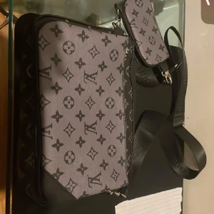 Trio axelväska monogram svart/grå - Snygg axelväska  i svart och grått med klassiskt monogrammönster. Väskan har justerbar axelrem, silverfärgade dragkedjor och en mindre avtagbar ficka framtill. Tillverkad i skinn med exklusiv känsla och stilren design.