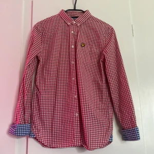 Rutig skjorta från Lyle & Scott Junior - Röd och vit rutig skjorta från Lyle & Scott Junior med klassisk krage och knappar framtill. Skjortan har långa ärmar med blåvitrutiga detaljer på insidan av manschetterna och bröstbrodyr med gul logga. Perfekt för dig som gillar färg och mönster.
