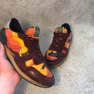 Valentino Rockrunner sneakers i röd camo - Färgstarka Valentino Rockrunner sneakers med camouflagemönster i rött, orange, gult och svart. Skorna har vinröda mockadetaljer, snörning och en klassisk gummisula. Perfekta för dig som vill sticka ut med exklusiva och unika sneakers.