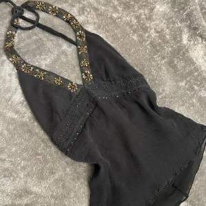 Svart halterneck topp med pärldetaljer - Supersnygg svart halterneck topp med guldiga pärlor och blommiga dekorationer längs ringningen. Tunn, luftig och lätt skrynklig textur. Perfekt för dig som vill sticka ut med coola detaljer och djup v-ringning. Öppen rygg!