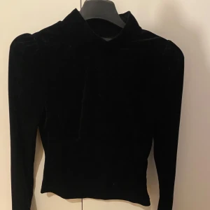 Svart sammetspolotröja med lång ärm - Snygg svart polotröja i mjuk sammet med lång ärm. Tröjan har en tight passform och hög krage som ger en stilren look. Perfekt för dig som gillar en klassisk men trendig vibe.