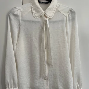 Vit blus med knyt och volangkrage Zara - Supersöt vit blus från Zara med lång ärm, volangdetaljer på kragen och knyt framtill. Blusen har en lätt glansig yta och klassisk passform, perfekt för dig som gillar romantiska detaljer och stilren look.
