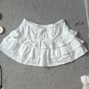 Vit volangkjol från Gina Tricot - Supersöt vit kortkjol från Gina Tricot med volanger och knytband i midjan. Kjolen har resår i midjan för extra komfort och är gjord i mjuk bomull. Perfekt för en fräsch och somrig look! Skulle säga den sitter bra på S med!