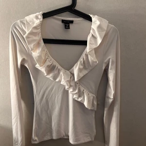 Vit volangtopp med v-ringning Amisu - Supersöt vit topp från Amisu med lång ärm och djup v-ringning dekorerad med volang. Tunn och mjuk känsla, perfekt för att lyfta en outfit med en romantisk touch. Enkel att matcha med både jeans och kjol.