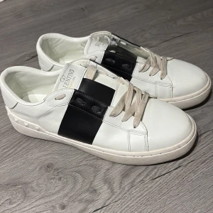 Valentino Garavani Open sneakers vit/svart - Snygga Valentino Garavani Open sneakers i vitt skinn med bred svart läderdetalj över mitten. Klassisk rund tå, platt sula och coola nitar längs hälen. Beige snörning och diskret logga på plösen. Perfekta för dig som gillar lyxig streetstyle. Pris kan diskuteras 