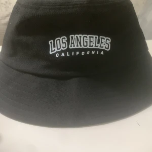 Svart bucket hat Los Angeles - Svart bucket hat med broderad vit text 'Los Angeles California' framtill. Klassisk form med mjuk brätte och avslappnad streetstyle-vibe. Perfekt accessoar för att lyfta din look med en touch av Kalifornien.