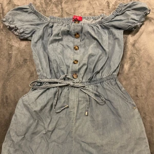 Blå offshoulder Romper från Guess - Supersöt blå kortklänning från Guess i jeansliknande bomullstyg. Offshoulder-modell med korta puffärmar, knappar framtill och knytskärp i midjan. Perfekt för varma dagar när du vill vara både chill och snygg.