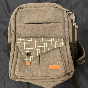 Grå ryggsäck från Leke Sky - Rymlig grå ryggsäck från Leke Sky med flera fack, USB-port på sidan där man själv lägger in en power bank i innerfickan och coolt mönstrad ficka framtill. Invändigt finns smarta fack och orange detaljer. Fack för bland annat laptop samt en nyckelhake.