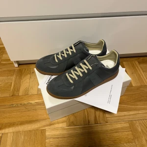 Grå Maison Margiela Gats sneakers - Maison Margiela sneakers i grått med beige snörning och gummisula. Ovandel i en mix av mocka och skinn, klassisk siluett och diskreta detaljer. Perfekta för dig som gillar stilrena och tidlösa sneakers med premiumkänsla. Har används en gång men inget som syns och kvitto från farfetch skickas med.