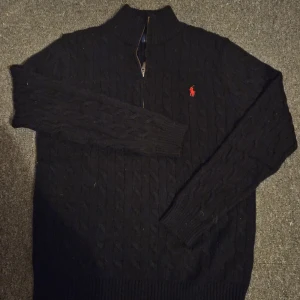 Svart stickad tröja Polo Ralph Lauren - Kabelstickad svart tröja från Polo Ralph Lauren med halv dragkedja och röd broderad logga på bröstet. Tröjan har hög krage och långa ärmar, perfekt för lager-på-lager. Klassisk och stilren design som funkar året runt.