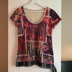 Färgglad blommig topp från Desigual - Unik topp från Desigual med färgstarkt blommigt mönster i rött, lila och orange. Framsidan har coola patchwork-detaljer och snörning, medan ryggen är ljus med broderad logga och spetsinsats upptill. Kortärmad och rund halsringning, perfekt för dig som gillar statement-plagg.